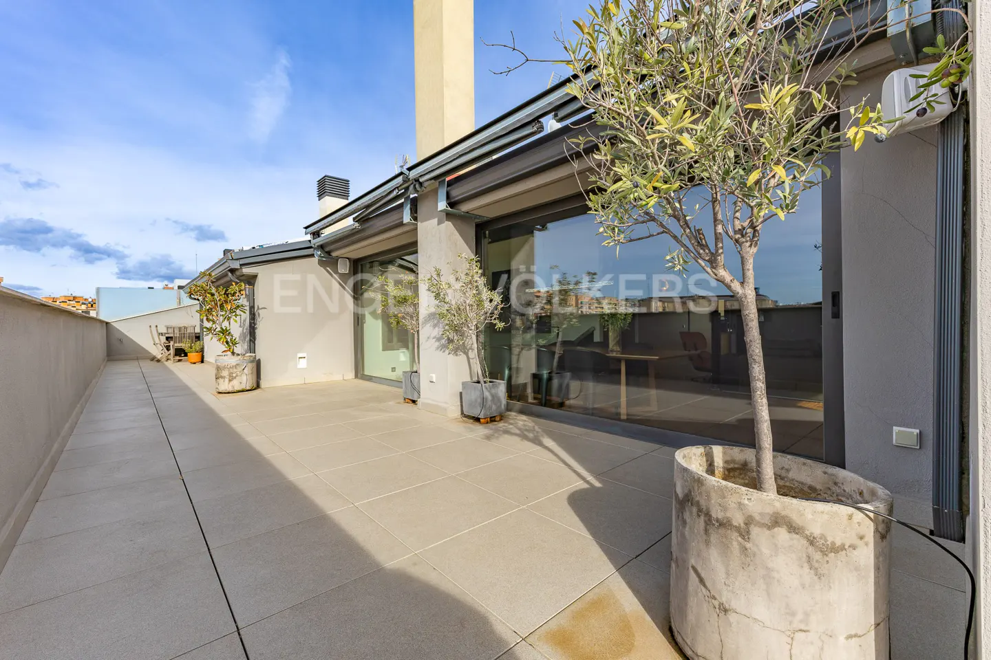A rooftop terrace with potted trees, gray tile flooring, and sliding glass doors. Blue sky is visible.