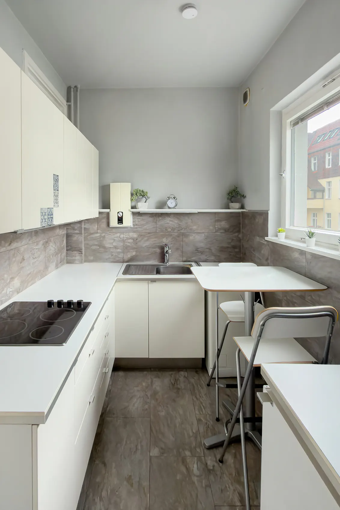 Bright kitchen with white cabinets, countertops, and a small table with two chairs. Gray backsplash and floor tiles. Window with a view of buildings.