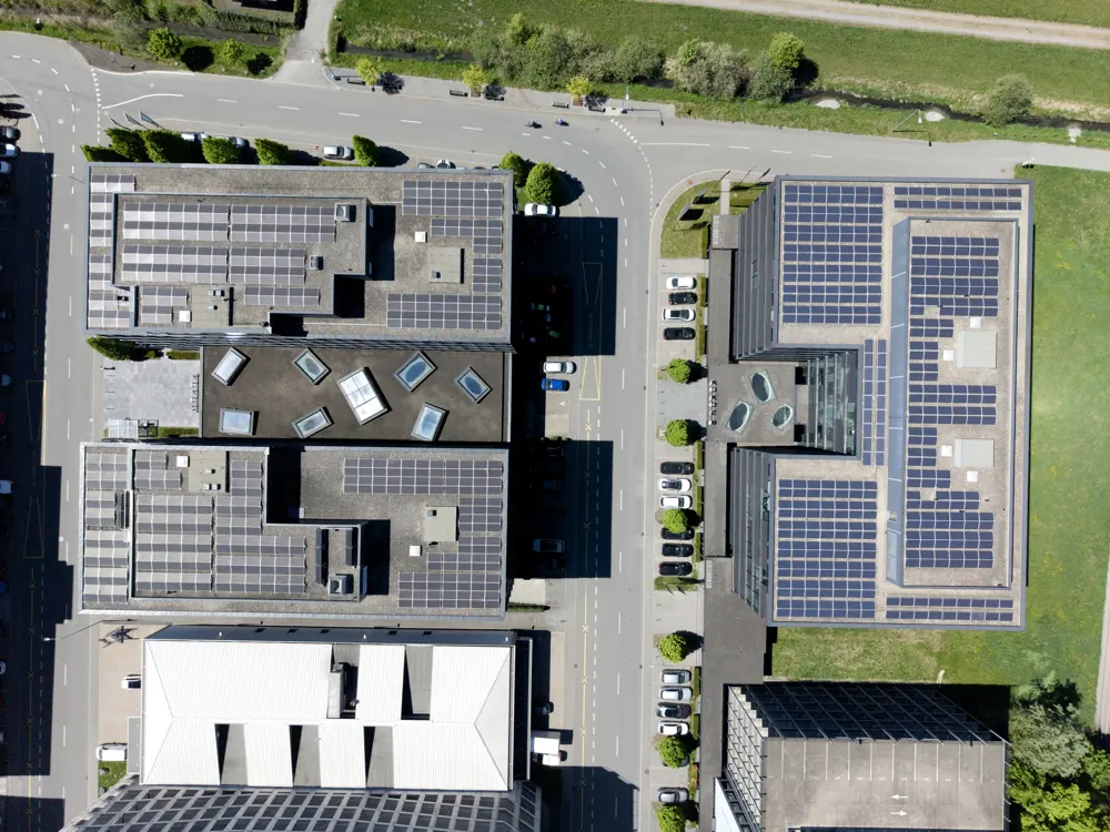 Aerial view of modern buildings with solar panels on roofs, cars parked along the street, and green spaces.