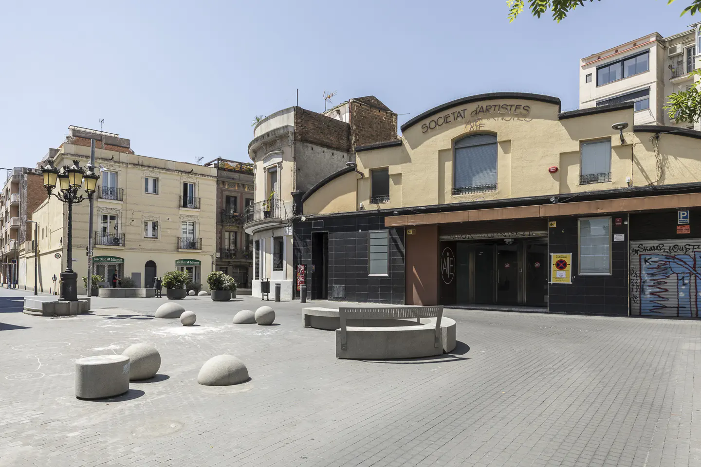 City square with stone benches and "Societat d'artistes" building. The square is paved with gray bricks and the sky is blue.