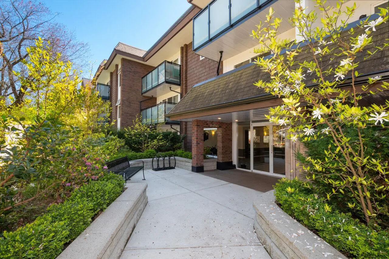 Exterior view of a brick apartment building with glass balconies, a covered entrance, and landscaped grounds.