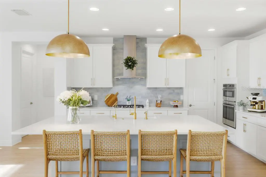 Bright, modern kitchen with white cabinets, a large island with wicker stools, and gold pendant lights. Stainless steel appliances are visible.