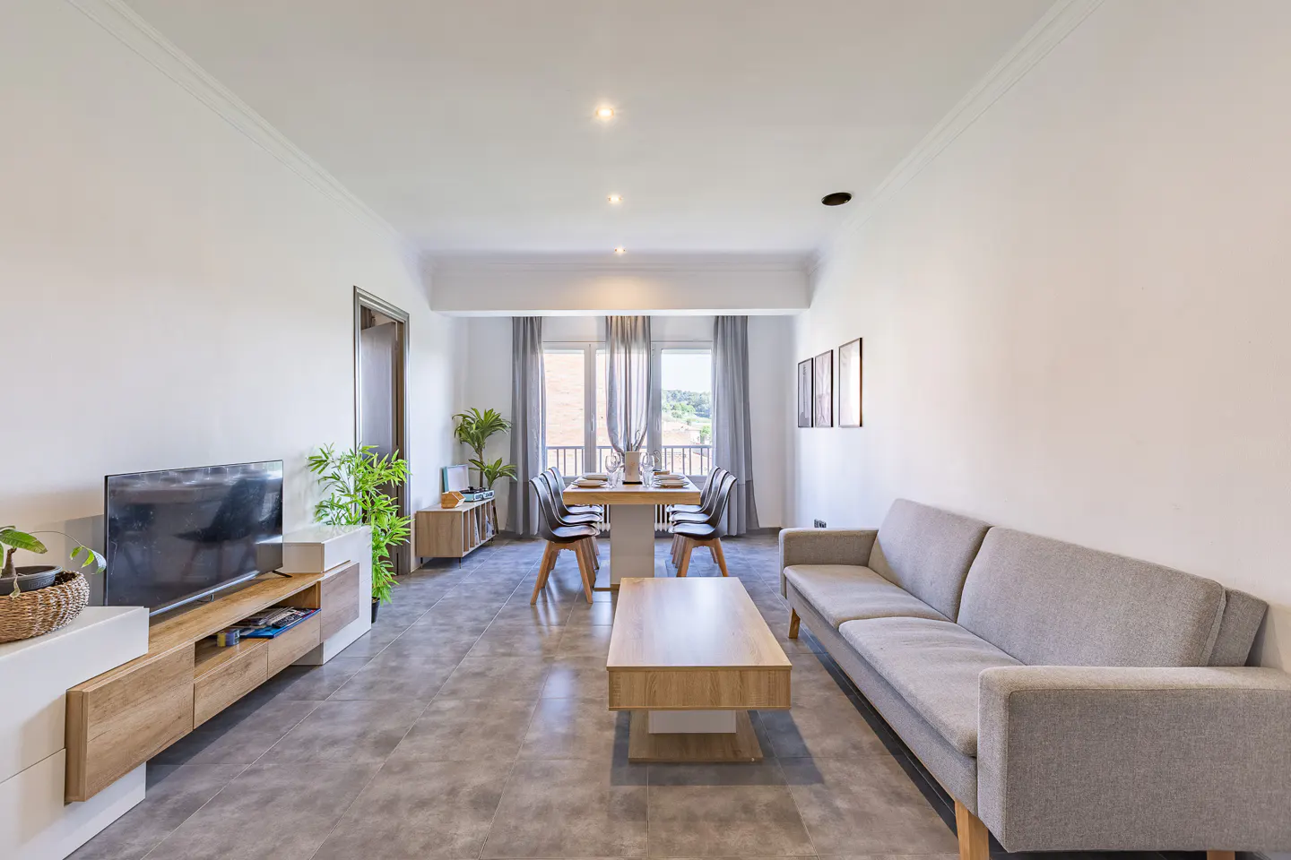 Bright living room with gray sofa, wood coffee table, TV, and dining table set for six near windows.