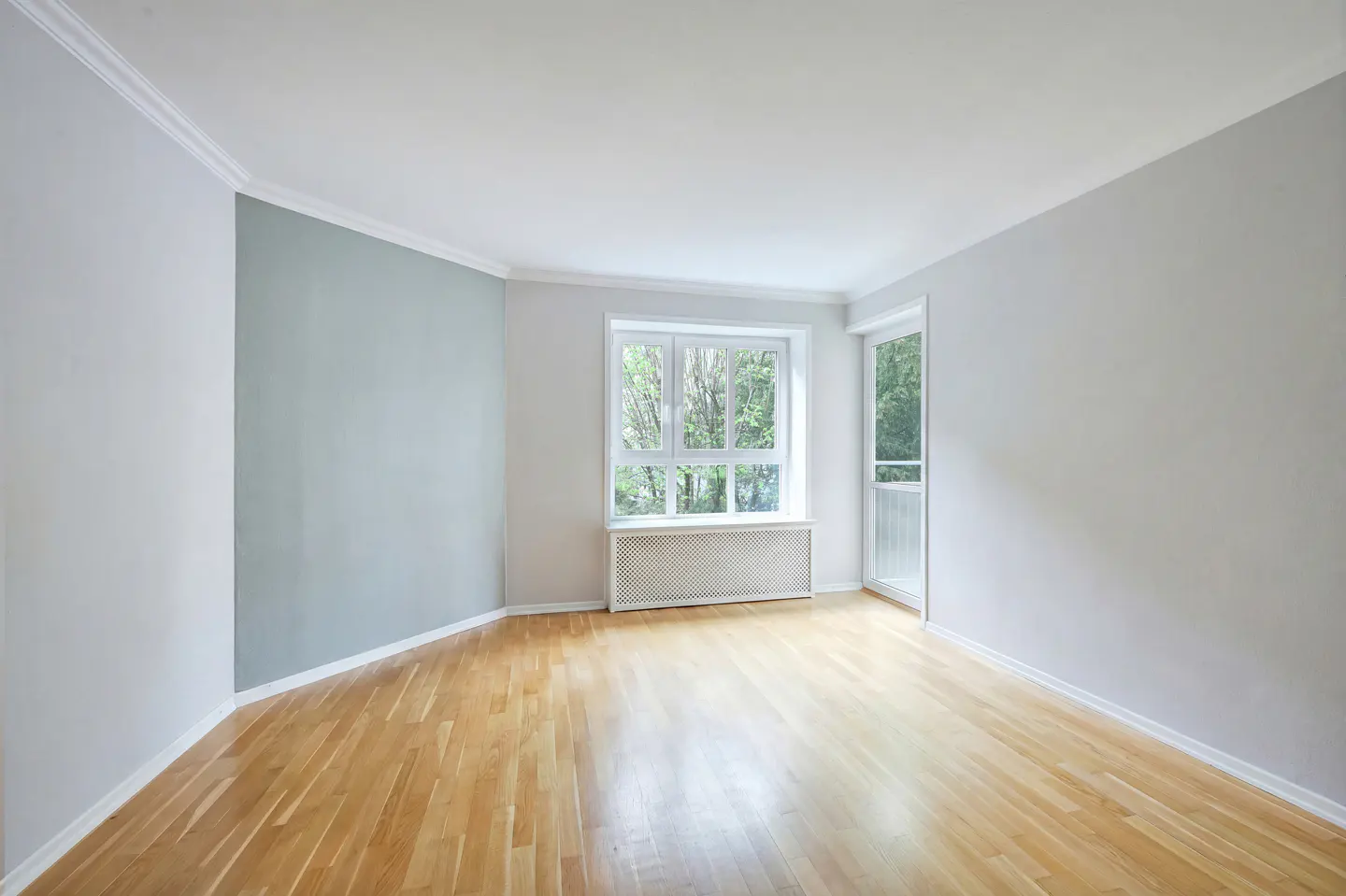 Bright, empty room with light wood floors, white walls, and a large window with a radiator. A glass door leads to a balcony.