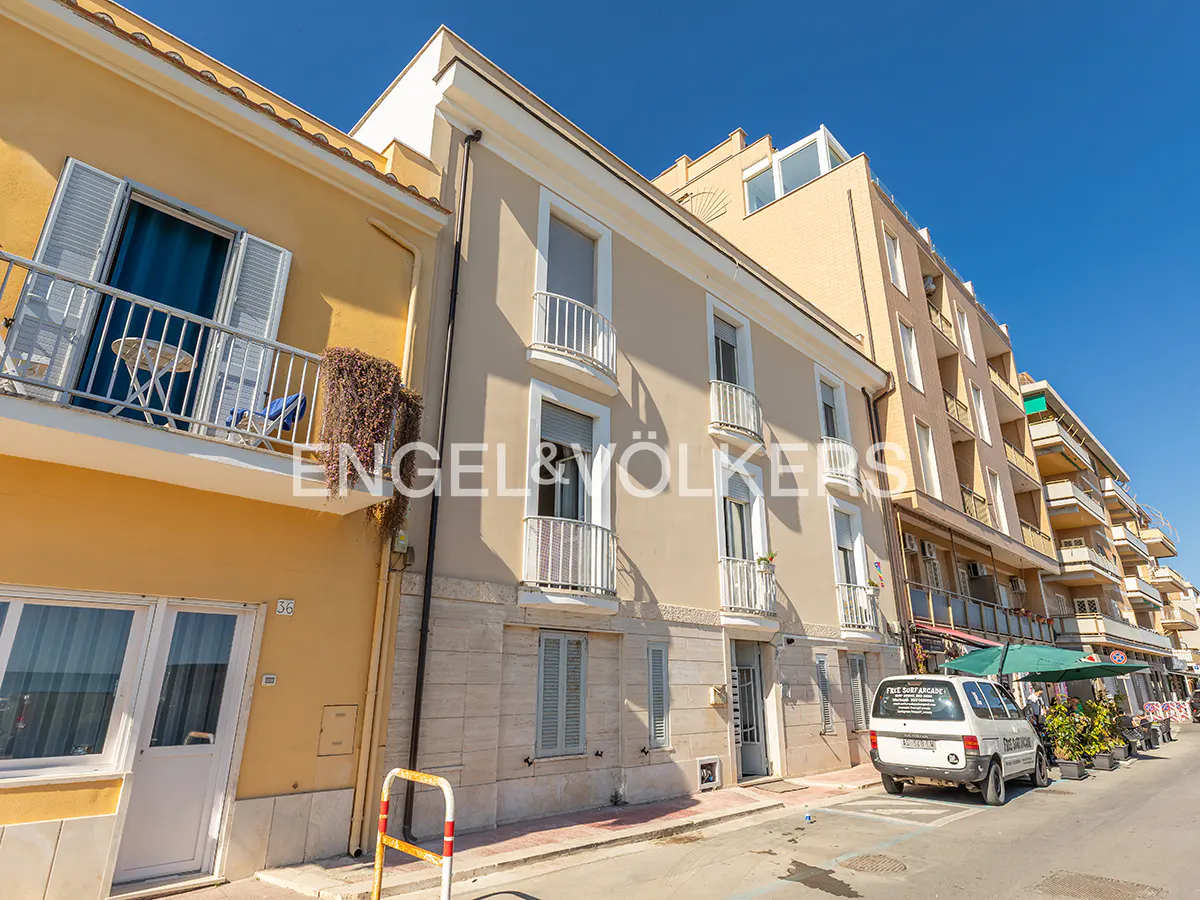 Exterior view of a multi-story building with balconies, a yellow building on the left, and a white van parked on the street.