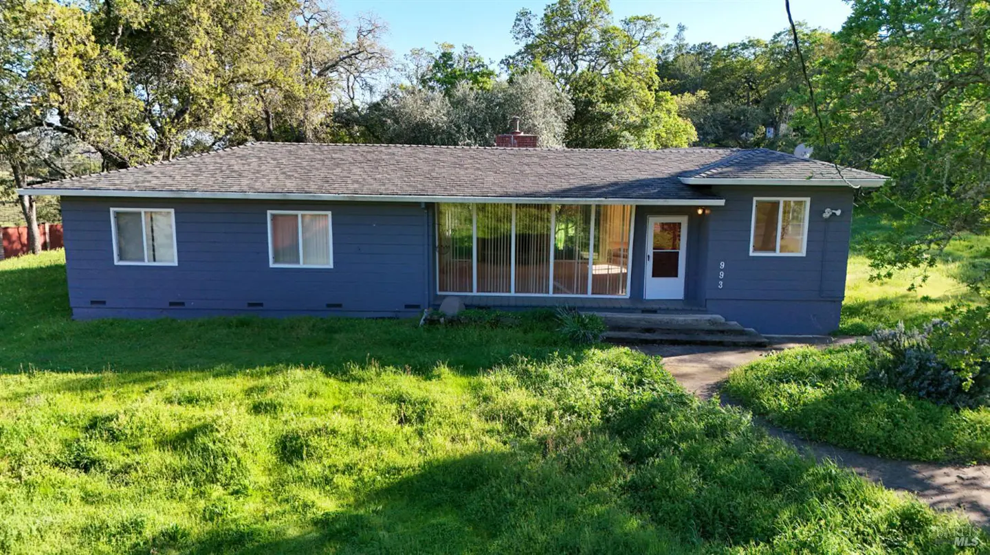 A single-story blue house with a gray roof, white trim, and a sunroom, surrounded by green grass and trees.