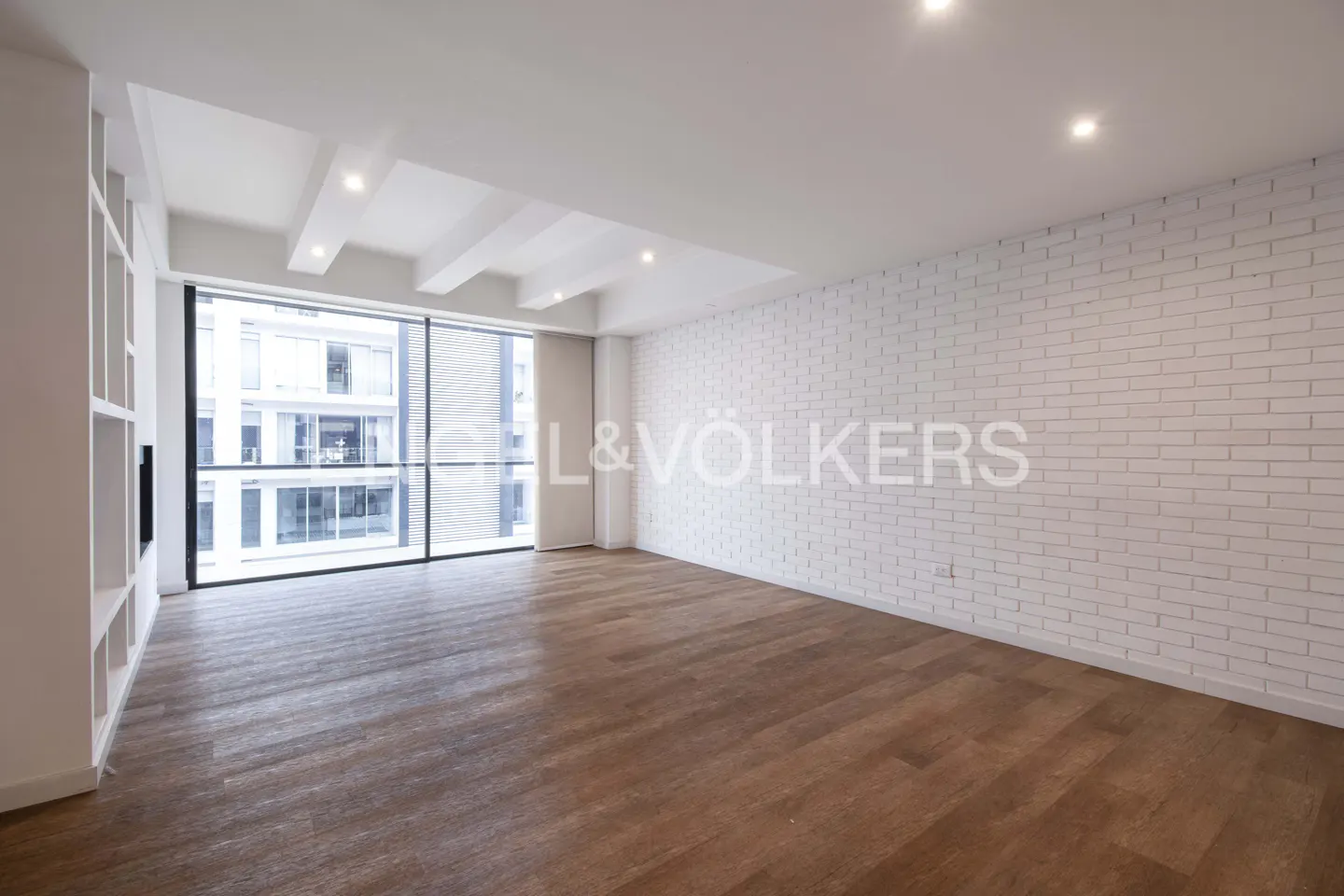 Bright, empty room with hardwood floors, white brick wall, and large window. Built-in shelves on the left. Engel & Völkers logo on the wall.