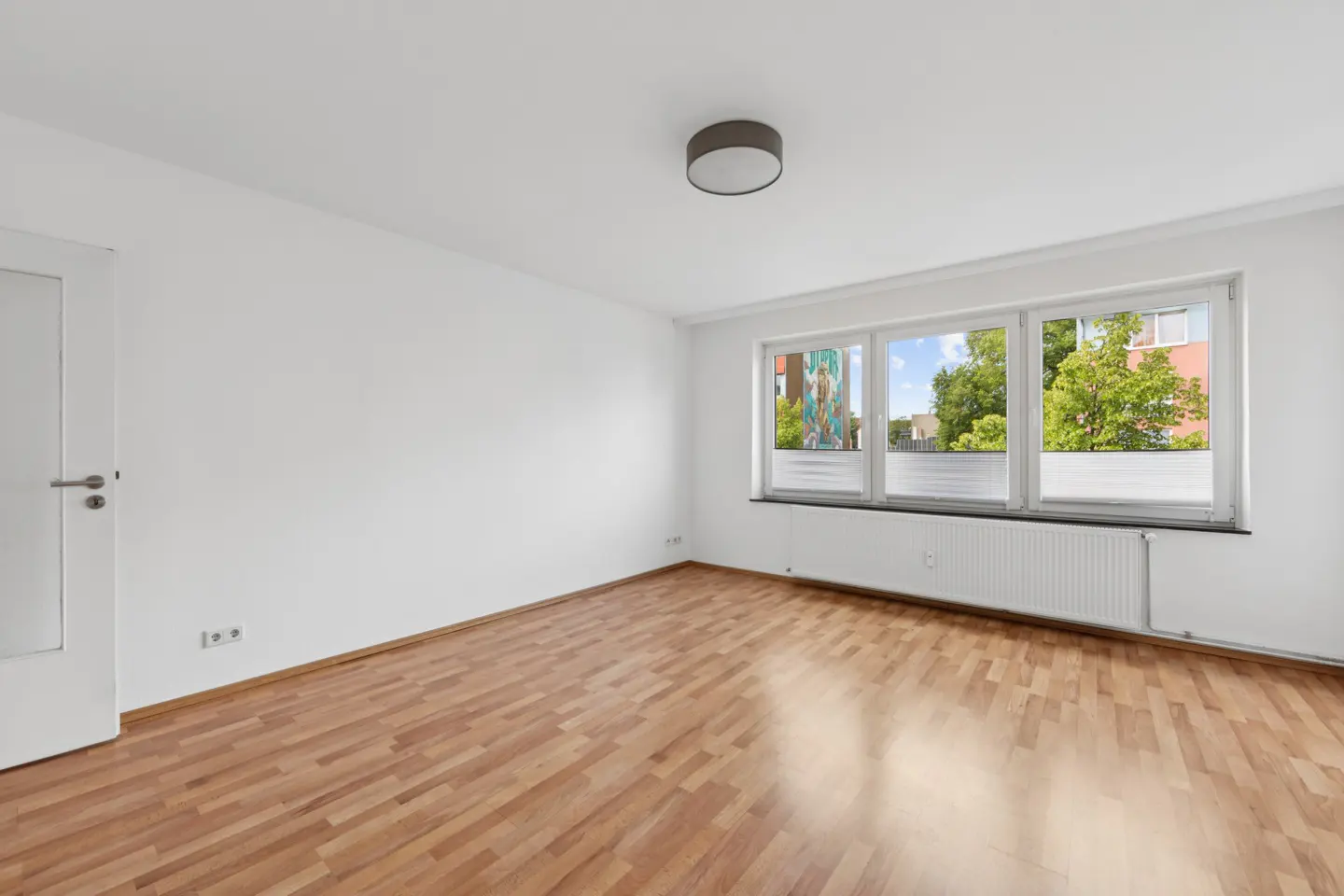 Bright, empty room with wood floors, white walls, and a large window showing trees and buildings outside.