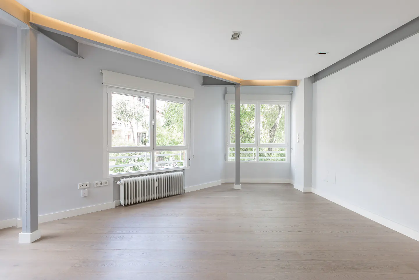 Empty, bright room with light wood floors, white walls, large windows, and exposed grey structural beams.