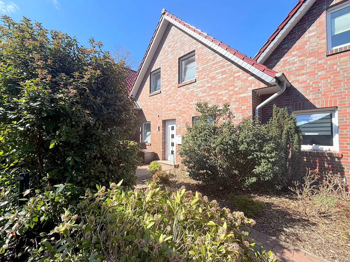 A two-story brick house with a red roof, white door, and green bushes in the front yard.