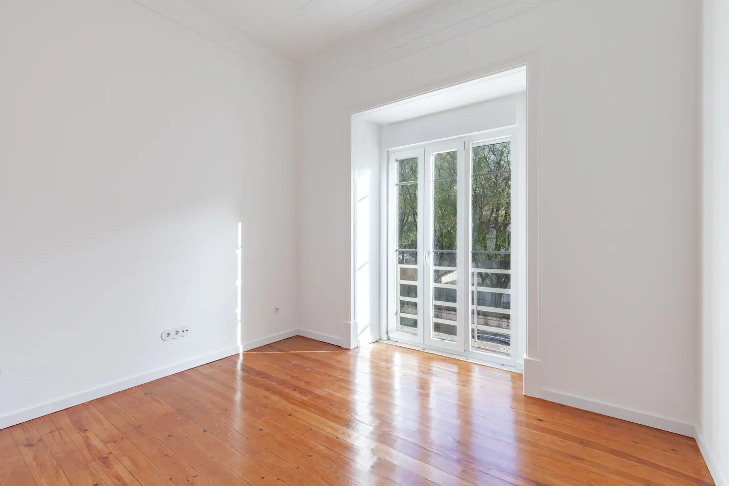Bright, empty room with white walls and a shiny wood floor. Double doors lead to a balcony with trees visible outside.