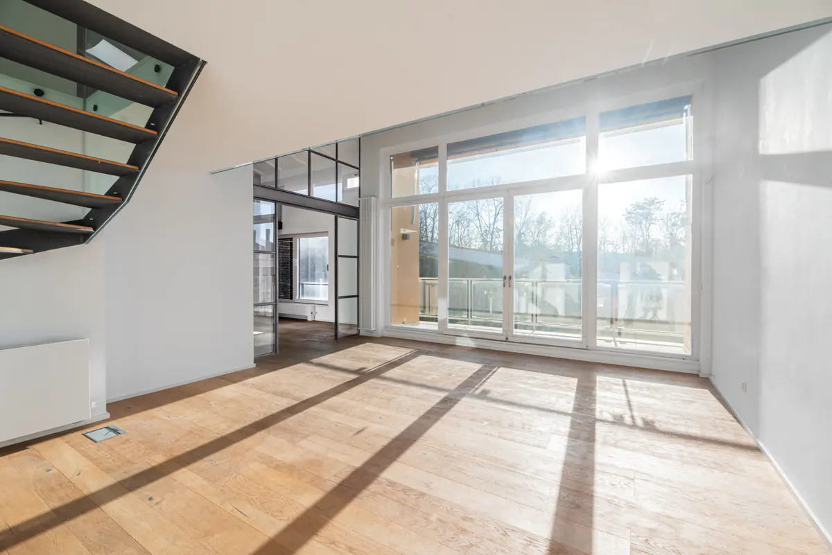 Bright, empty room with wood floors, white walls, and large windows with a balcony. A metal staircase is on the left.