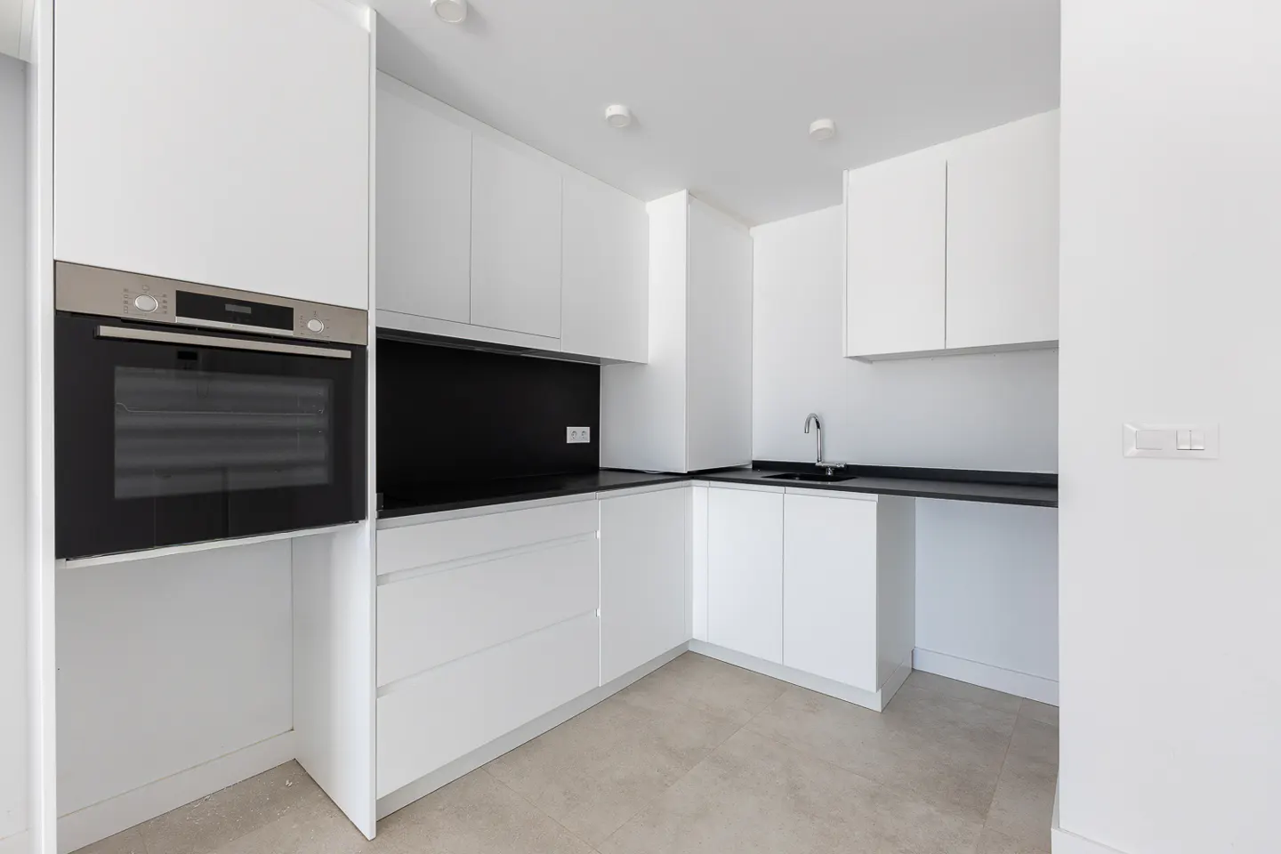 Modern kitchen with white cabinets, black countertops, a built-in oven, and light tile flooring.