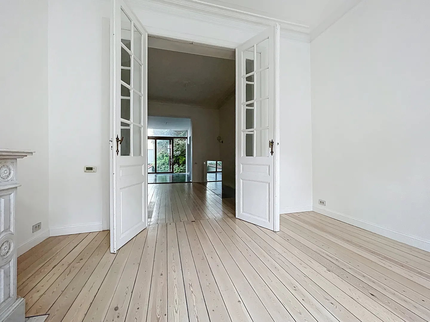 Bright, empty room with light wood floors and white walls. Open, white French doors lead to another room with a glass door to the outside.