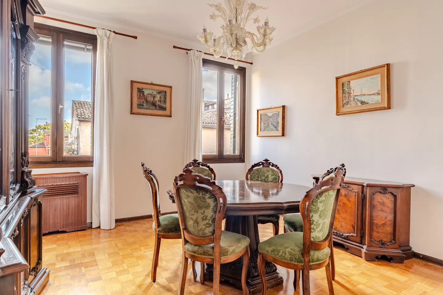 A dining room with a dark wood table, green chairs, and a crystal chandelier. Windows show buildings outside. Paintings adorn the white walls.