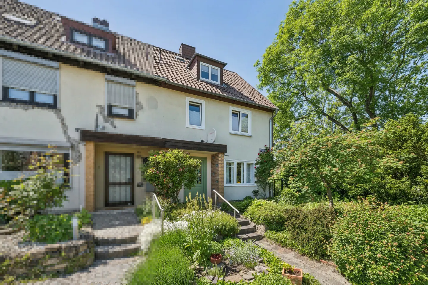 Two-story beige townhouse with a brown tiled roof, green front door, and a lush, landscaped garden under a clear blue sky.