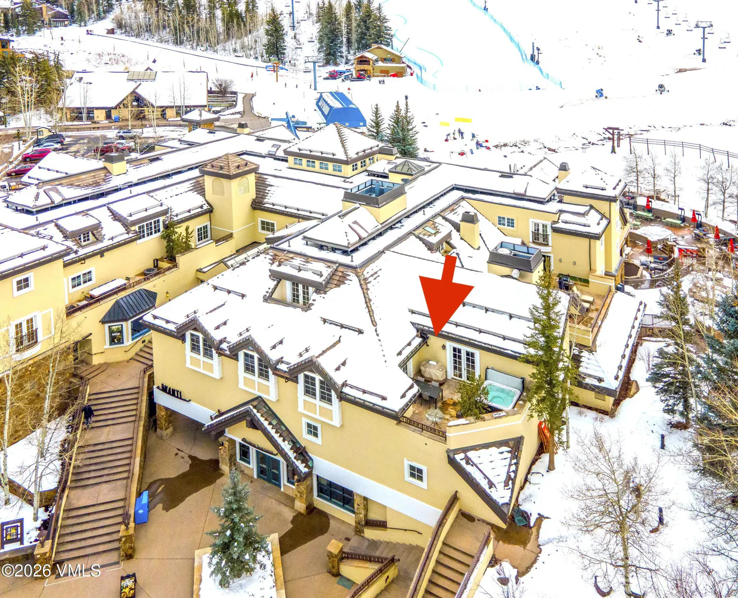 Aerial view of a tan ski resort building with snow-covered roofs, a hot tub on a deck, and a red arrow pointing to a unit.