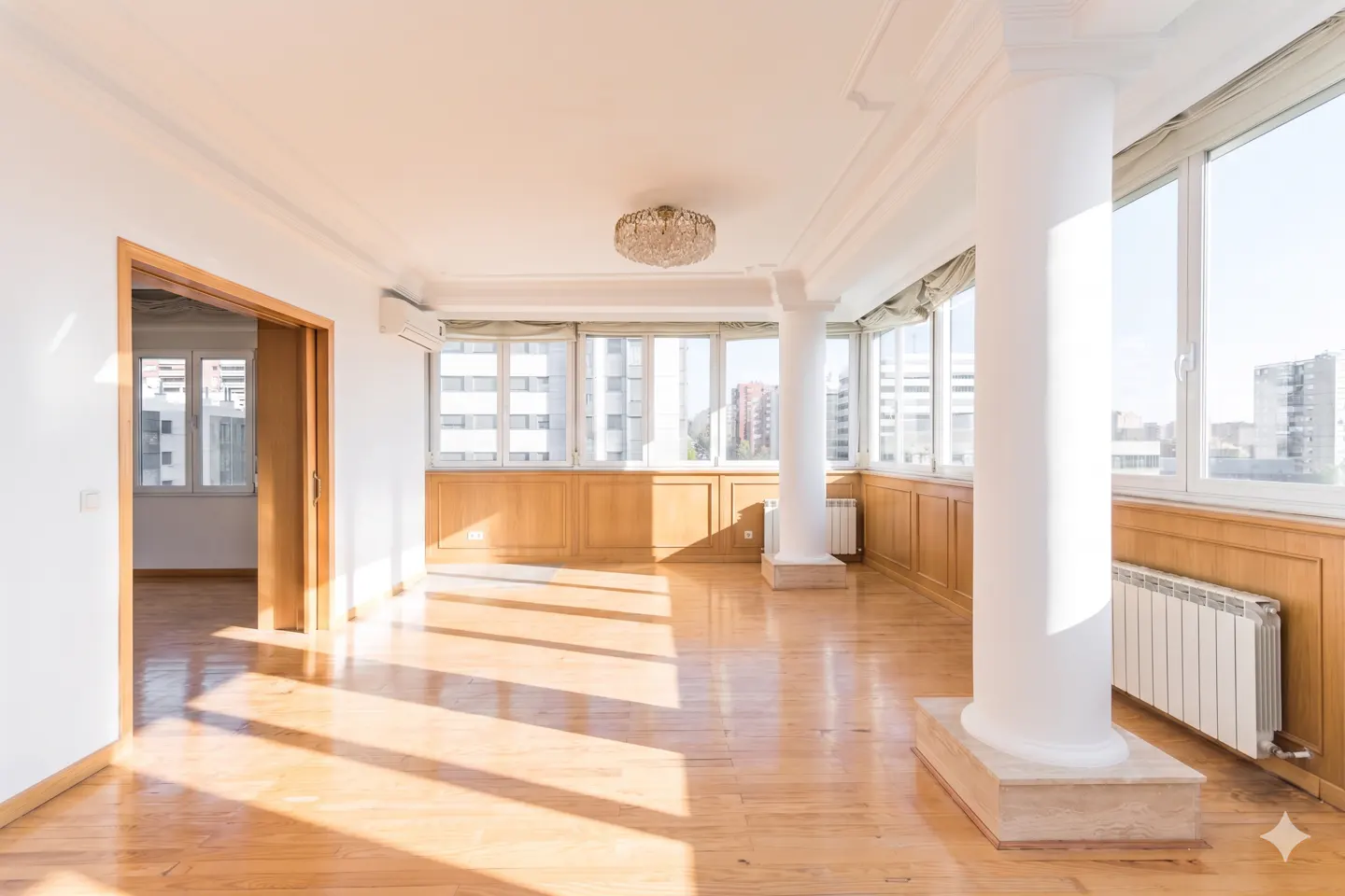 Bright, empty room with wood floors, white walls, and large windows. A white column stands near the windows. A chandelier hangs from the ceiling.