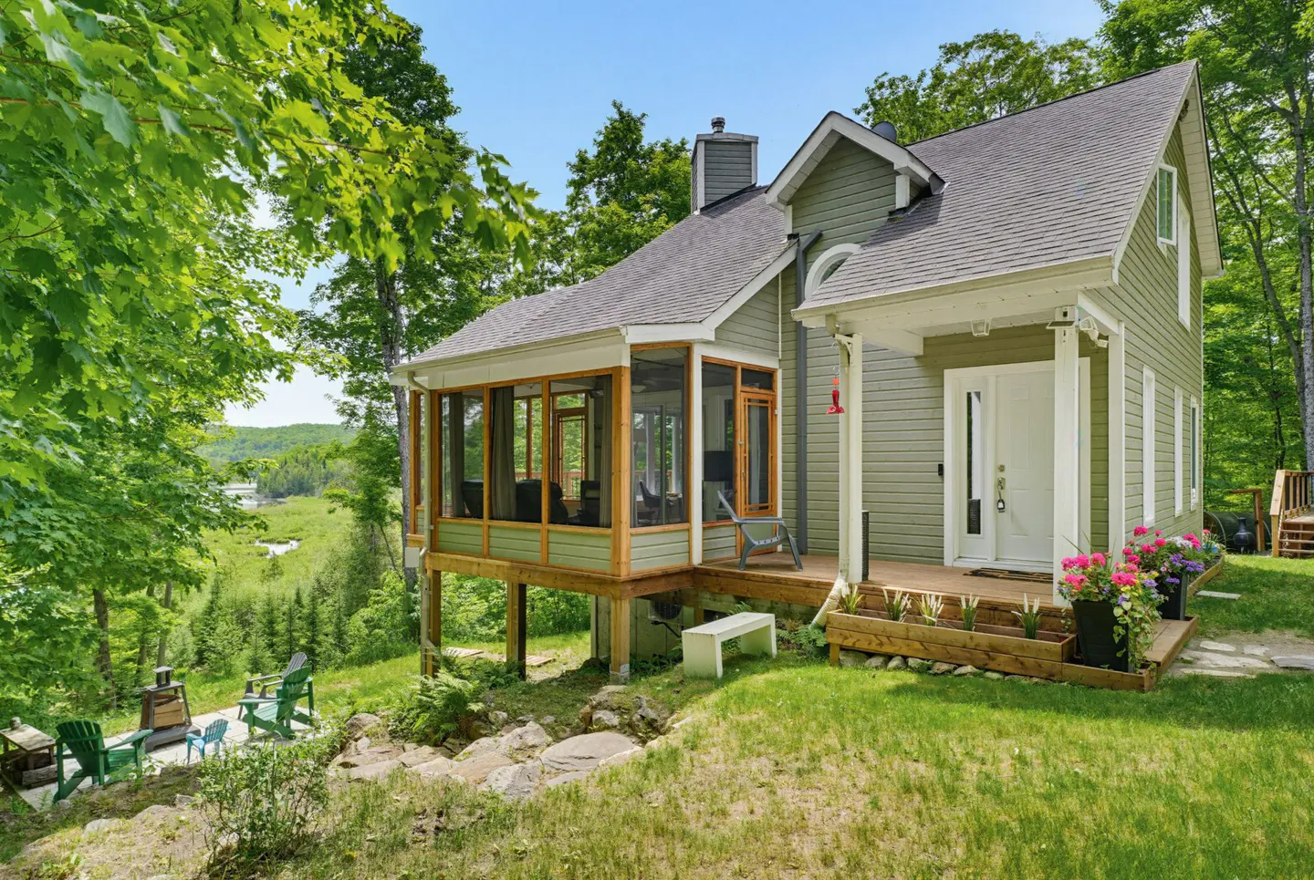 Sage green cottage with a screened porch, white front door, and wooden deck, surrounded by lush trees and a grassy yard.