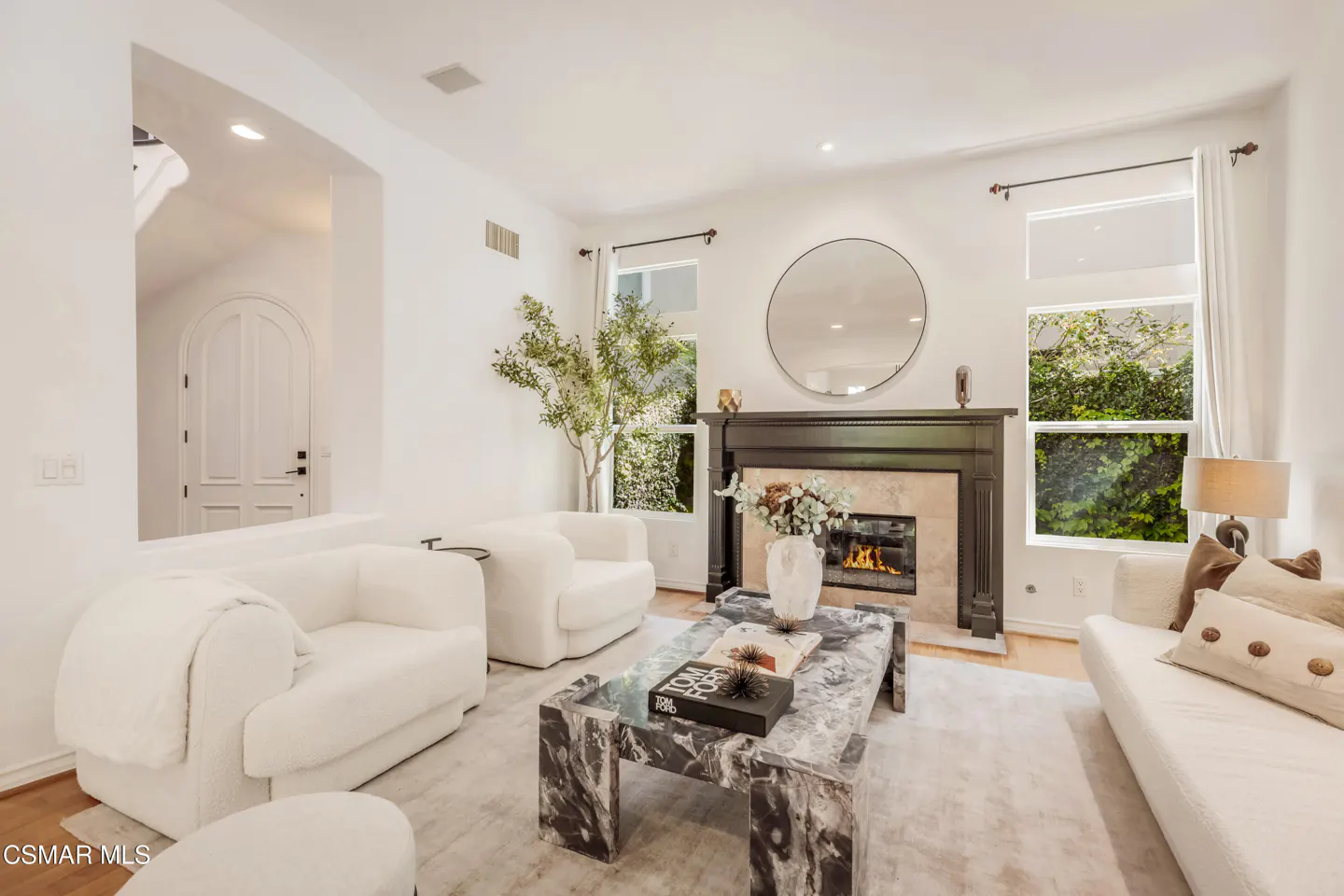 Bright living room with white walls, white sofas, and a marble coffee table. A fireplace with a round mirror above it is centered on the back wall.
