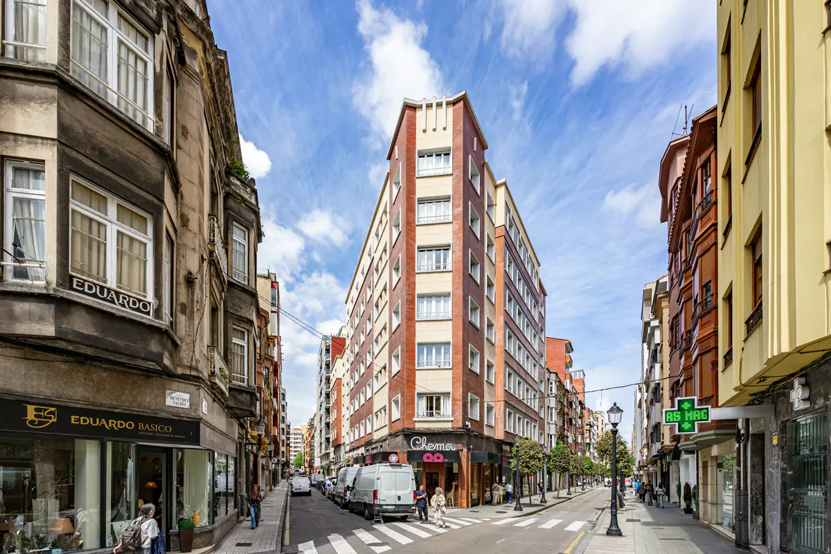 A narrow city street with a tall, wedge-shaped brick building at the center, flanked by historic buildings under a blue sky.