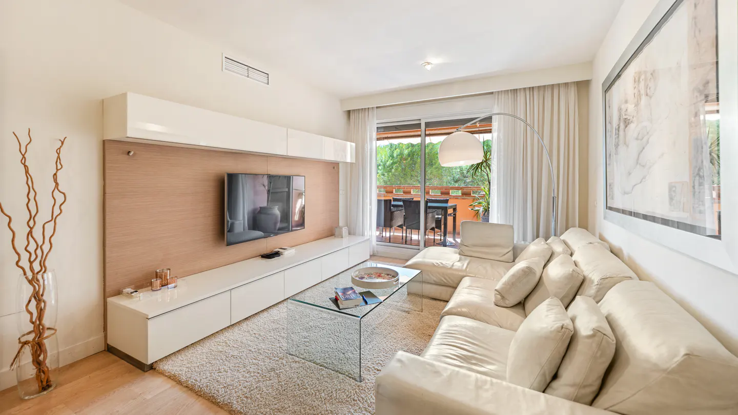Bright living room with white sofa, glass table, and TV. Balcony with outdoor furniture visible through sheer curtains. Modern decor.