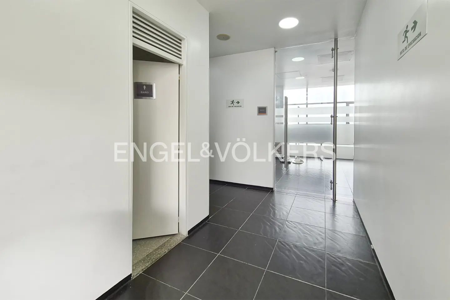 Hallway with white walls, dark tile floor, and a bathroom door on the left. A glass door leads to a bright office space.