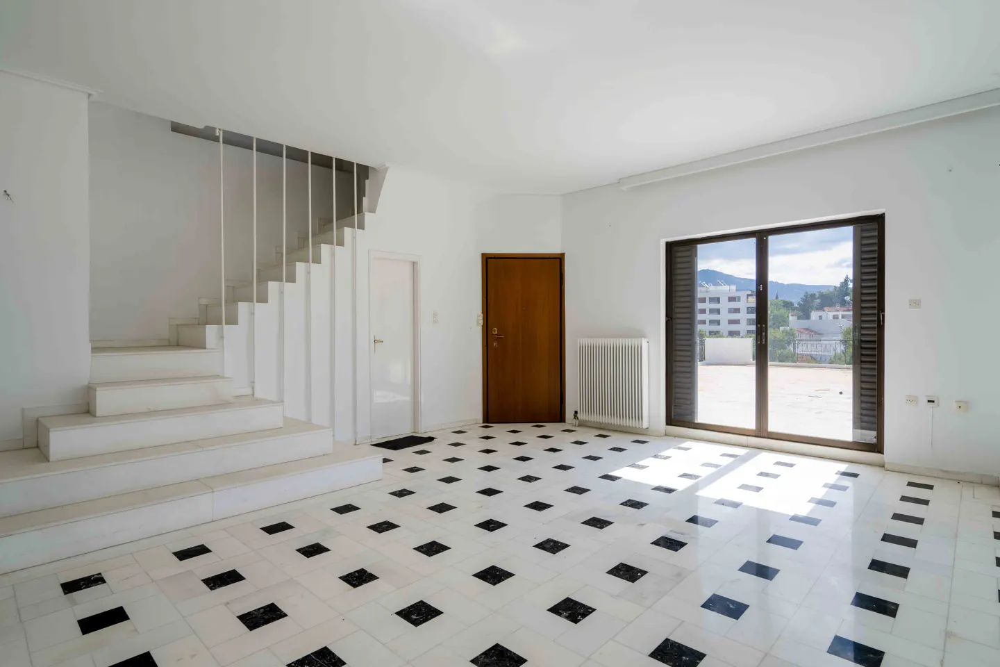 Bright, empty room with white walls, black and white tiled floor, stairs, and a door to a balcony with a view.