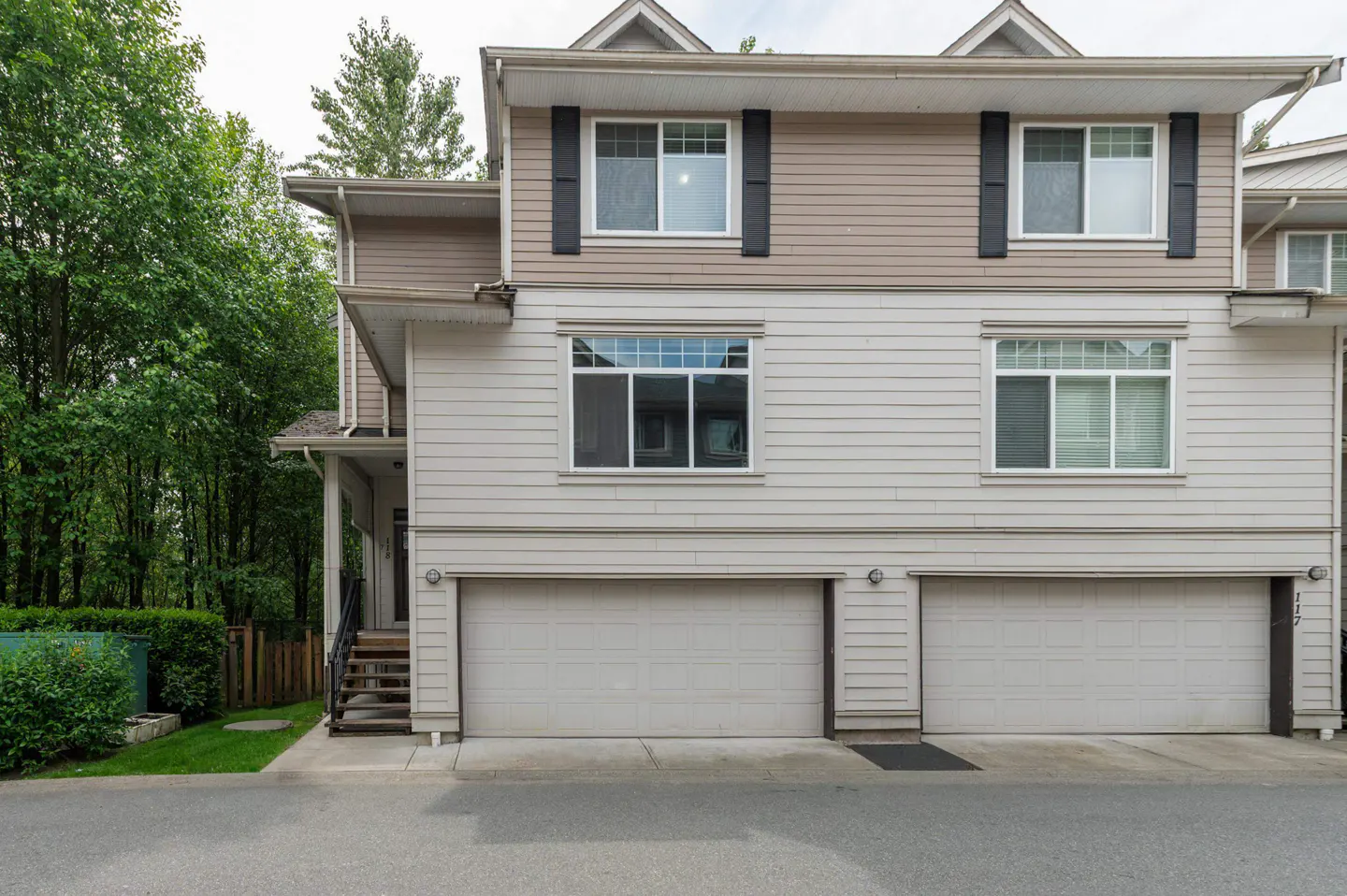 Exterior view of a two-story townhouse with beige siding, two garage doors, and a side entrance with stairs.
