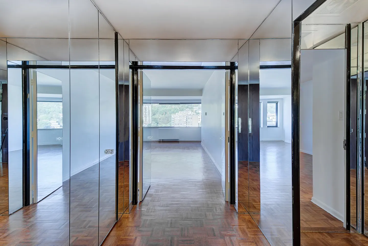 Hallway with parquet wood floors and floor-to-ceiling mirrored walls leading to a bright room with city views.