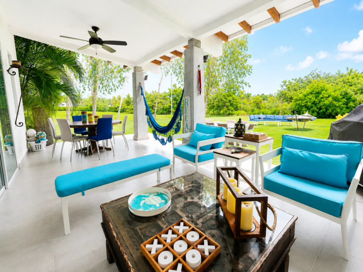 Covered patio with blue cushions, hammock, and tic-tac-toe game on a coffee table. Green lawn and trees in the background.