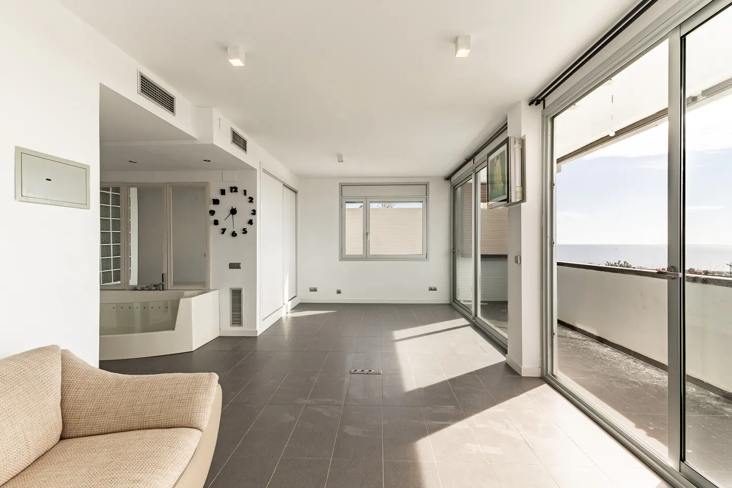 Bright, modern living room with gray tile floor, white walls, and large sliding glass doors to a balcony with ocean view. Beige sofa in foreground.