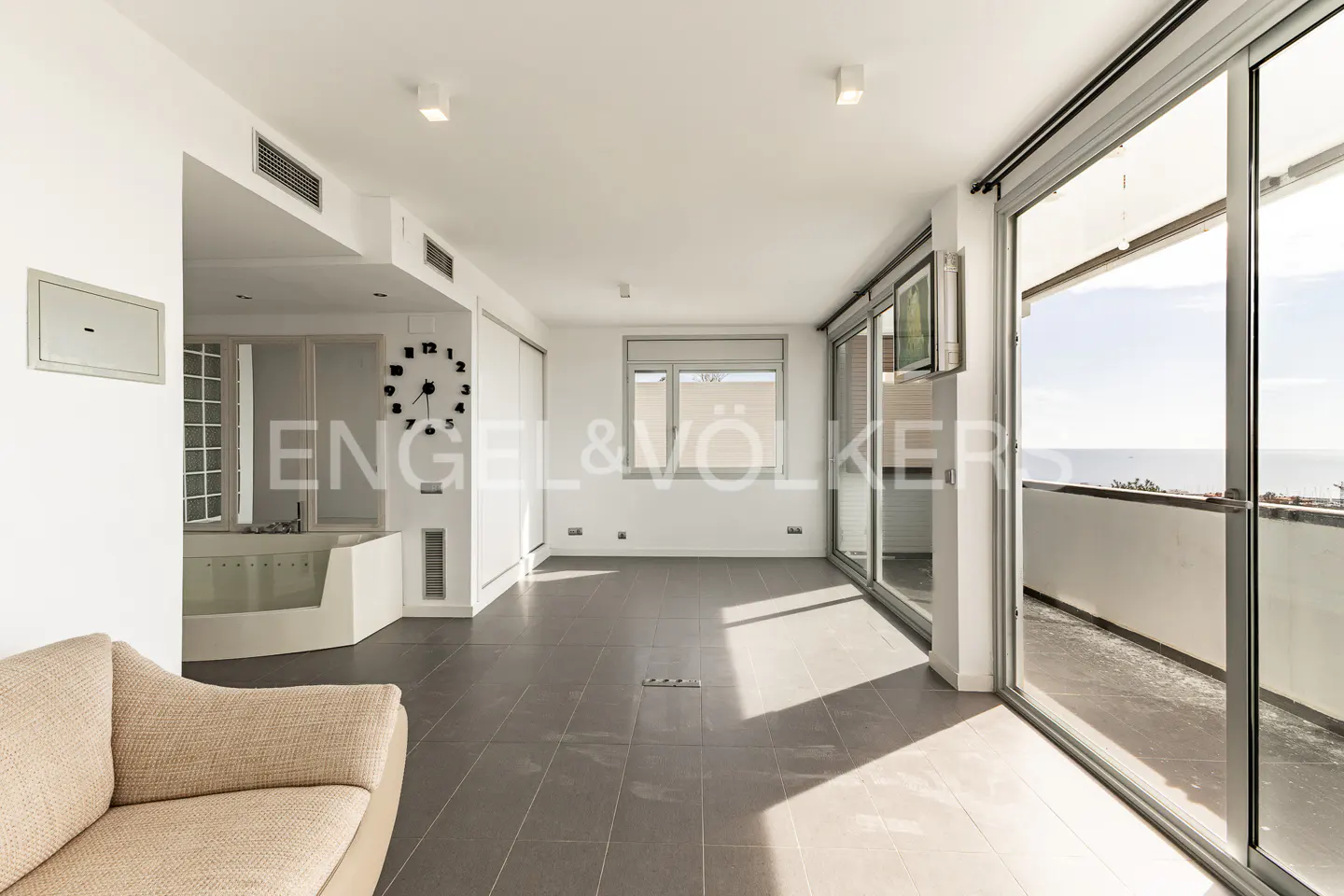Bright, modern living room with gray tile floor, white walls, and large sliding glass doors to a balcony with ocean view. Beige sofa in foreground.