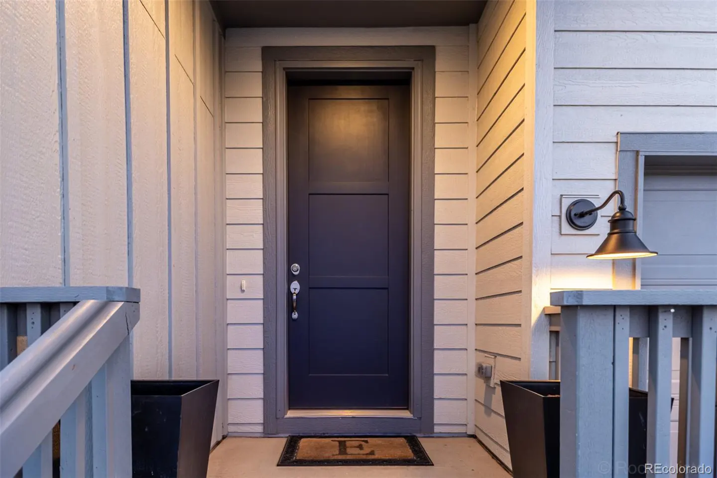 Exterior shot of a blue front door with gray trim, a welcome mat, and light-colored siding.