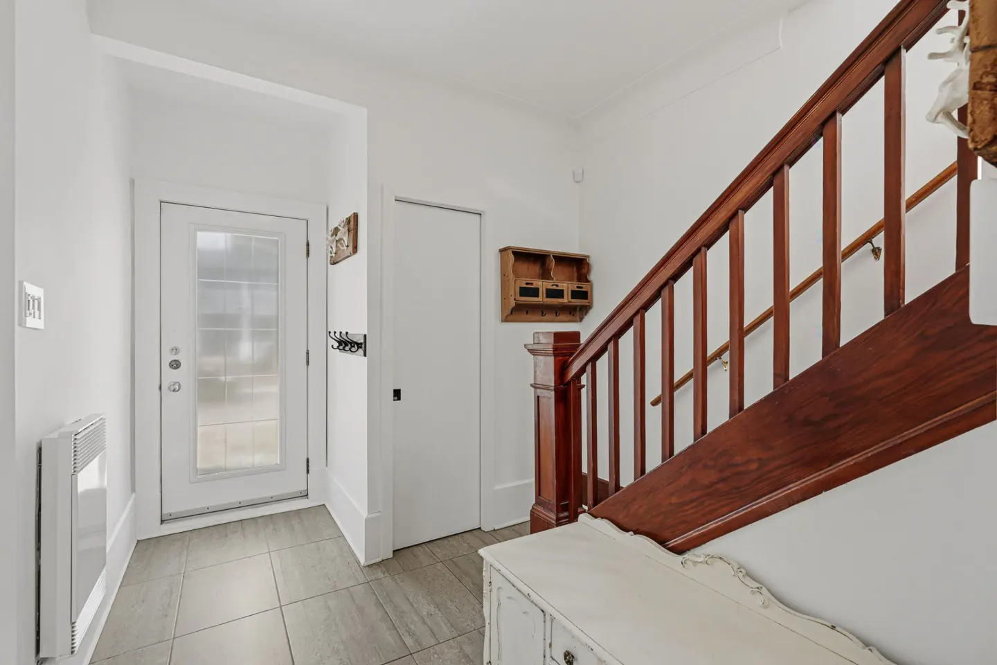 Bright entryway with white walls, gray tile floor, and a white door with glass panels. A dark wood staircase is on the right.
