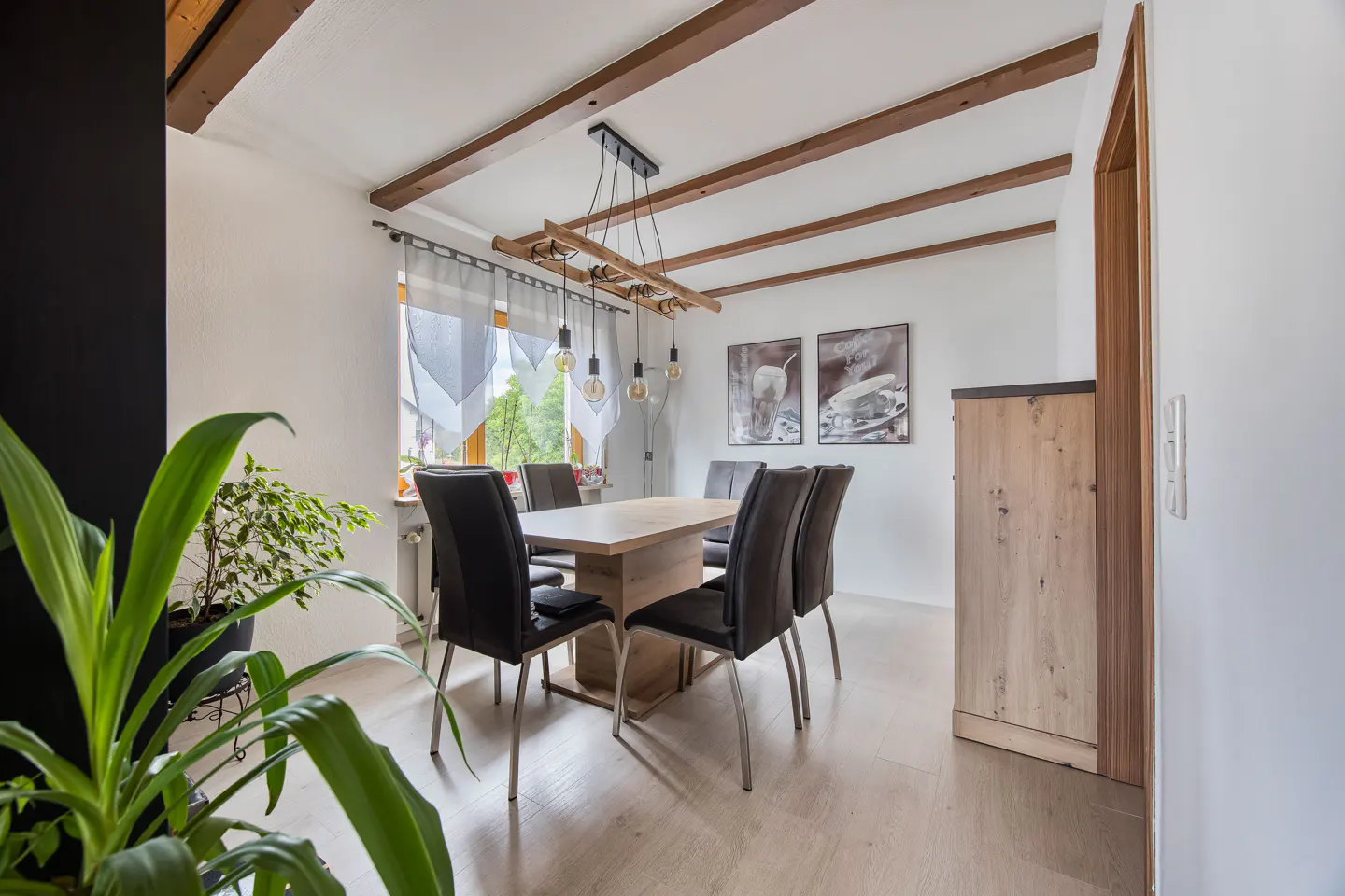 Dining room with a light wood table, black chairs, and a unique light fixture. Exposed beams on the ceiling and plants add warmth.