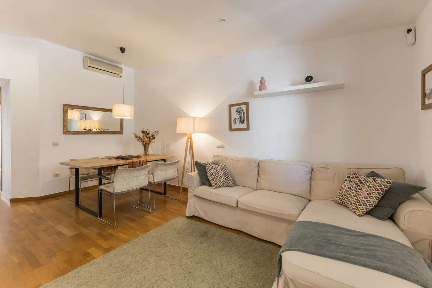 Bright living room with wood floors, white walls, and a beige sectional sofa. A dining table with four chairs sits near a mirror.