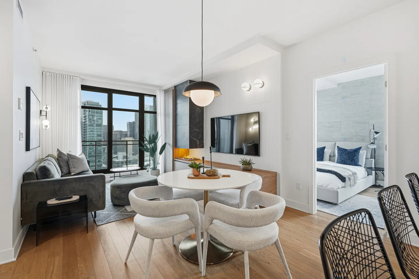 Bright, modern apartment interior with a gray sofa, white dining table, and a bedroom visible through an open doorway. City view from the window.