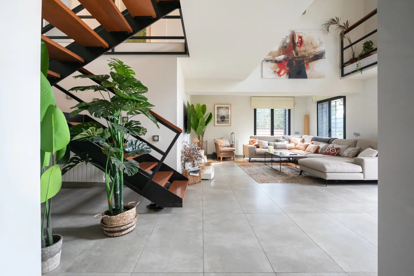 Modern open-plan living room with a black metal staircase, large potted plants, and a beige sectional sofa on tile floors.