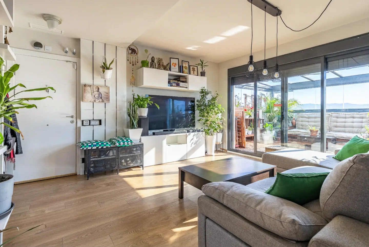 Bright living room with a gray sofa, wood floors, and a sliding glass door to a plant-filled balcony. A TV sits above a white cabinet.
