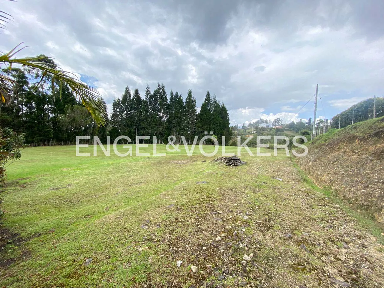 A large, green, grassy field with trees and a cloudy sky. The Engel & Völkers logo is superimposed on the image.