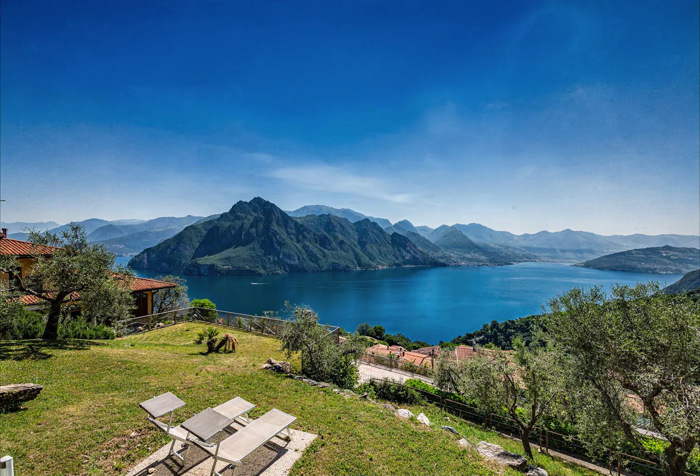 Scenic view of Lake Iseo, Italy, with mountains in the background. A green lawn with lounge chairs is in the foreground.
