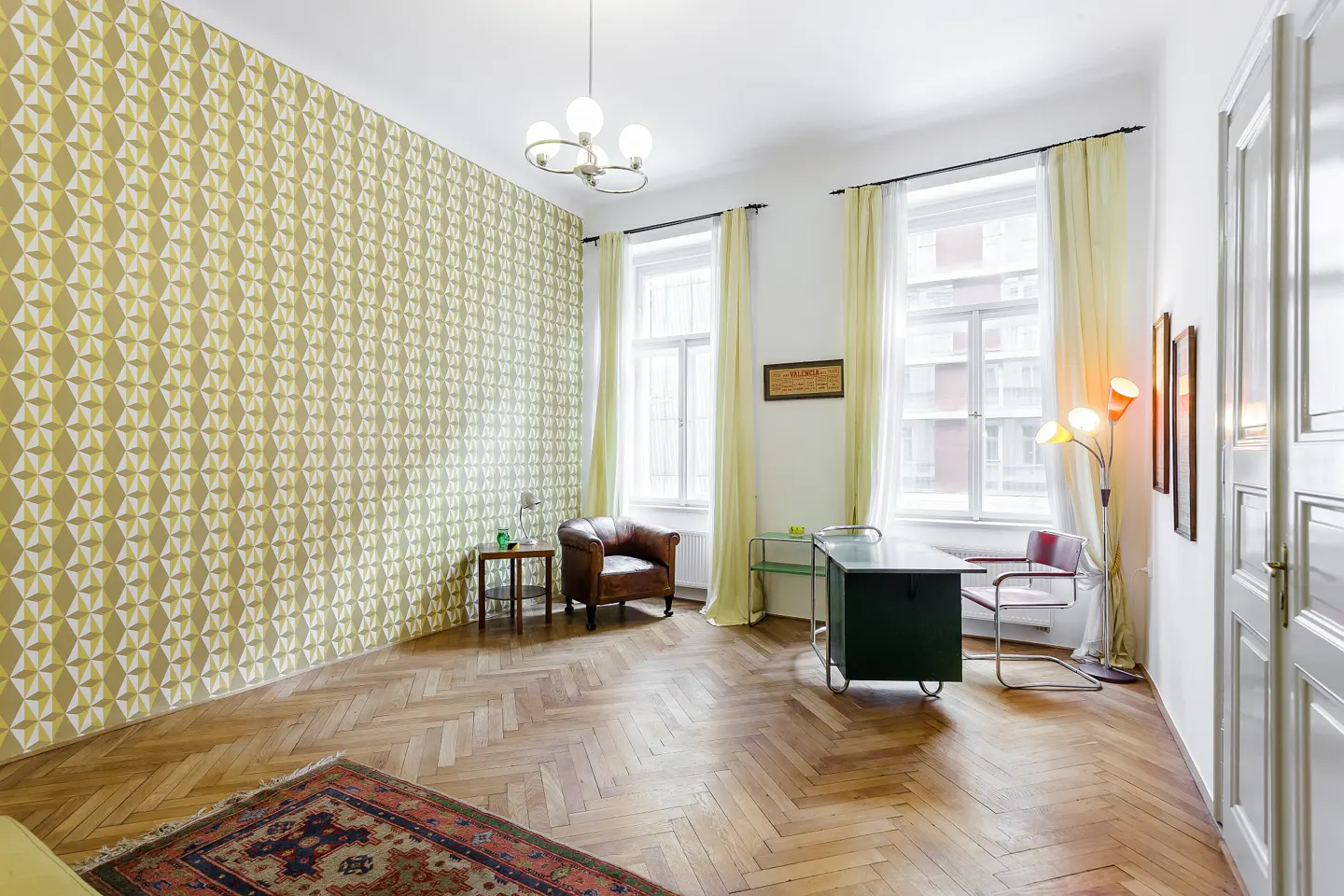 A bright room with parquet floors, patterned wallpaper, and a vintage desk. Natural light streams through the windows.