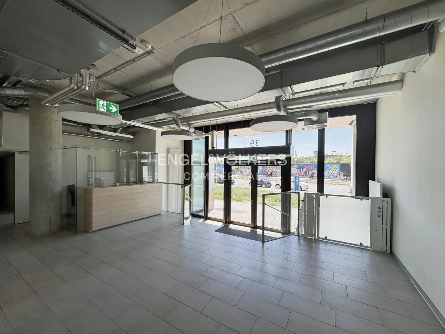 Empty commercial space with gray tile floor, exposed ductwork, and round pendant lights. Glass doors lead to a street view.