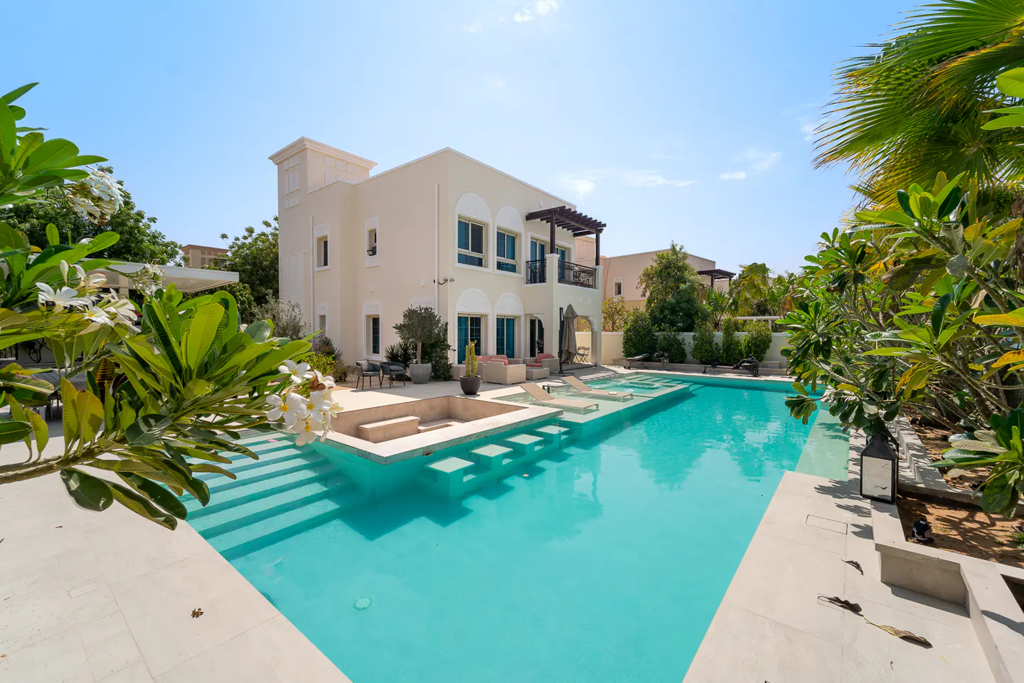 Exterior of a two-story beige house with a turquoise swimming pool and green foliage.