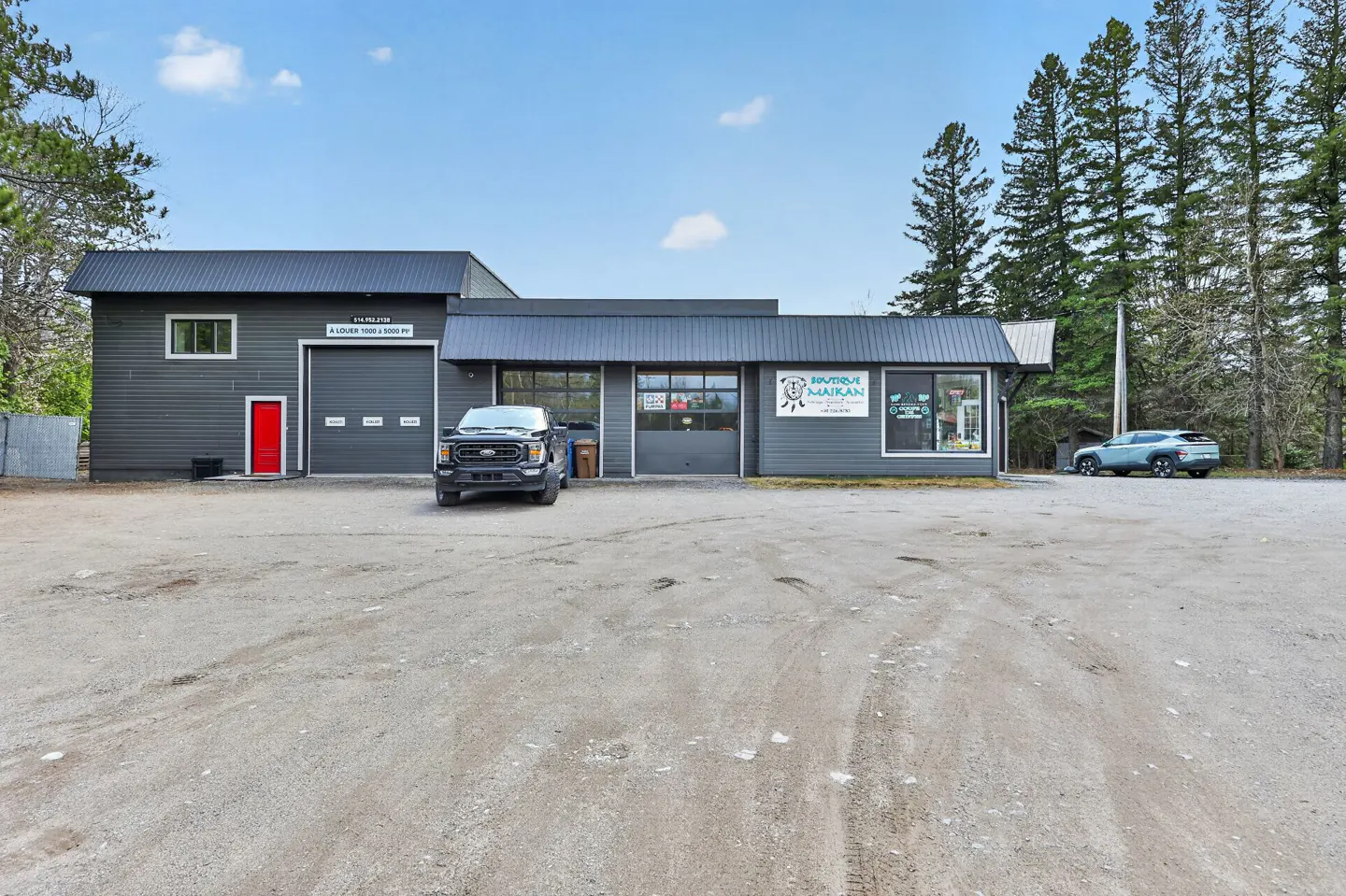 Gray commercial building with a red door and garage doors. A black truck is parked in front, and trees are in the background.