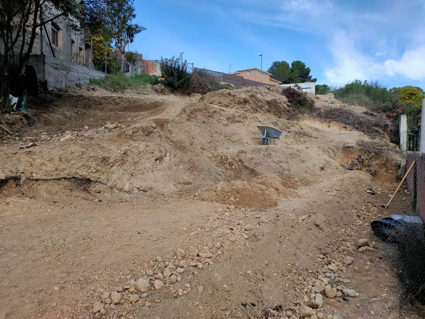 A sloped lot with exposed dirt and rocks, a wheelbarrow, and a few trees under a blue sky.