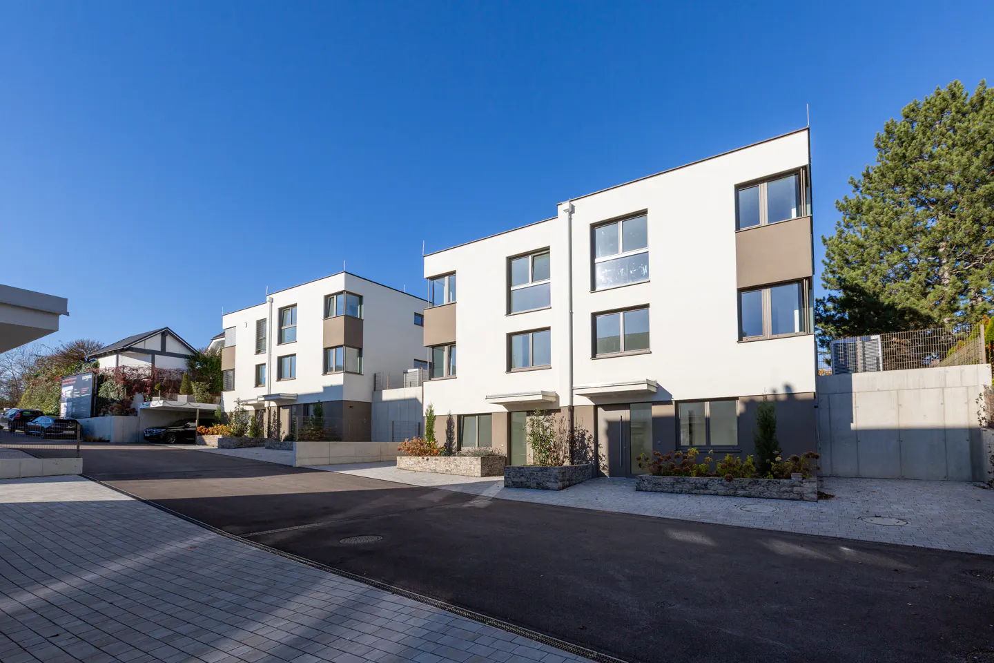Modern white townhouses with brown accents under a clear blue sky. A paved driveway leads to the homes, with landscaping in front.