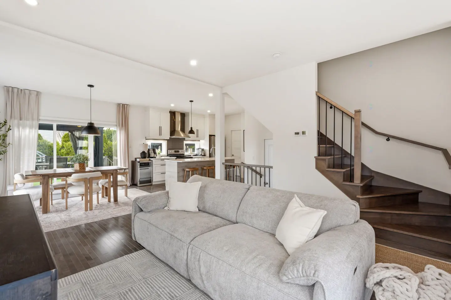 Open-concept living space with a gray sofa, white pillows, dining table, and kitchen. Stairs lead to the second floor.