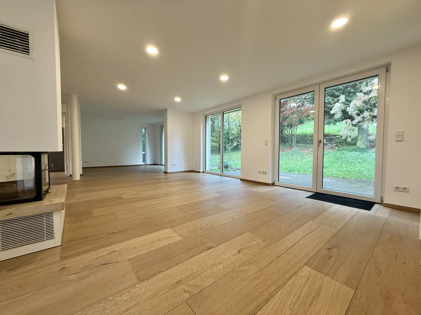 Bright, empty living room with light wood floors, white walls, and a fireplace. Sliding glass doors lead to a green yard.